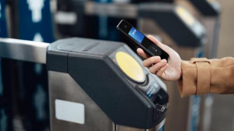 A person holding a mobile wallet up to a ticket barrier, either at a train station or a subway