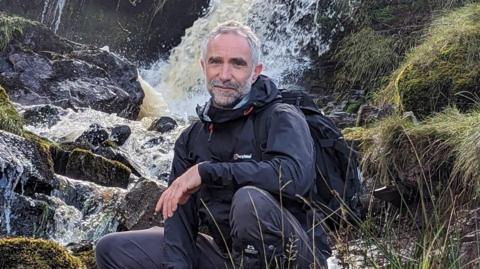 James Kirby, a British aid worker who was killed in Gaza. He is seen in a black mac and dark trousers crouching in front of a waterfall on a hillside. 