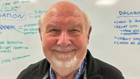 A smiling elderly man with white hair on the sides of his head and a short white beard. He is standing in front of a whiteboard with things written on it in blue and green ink.