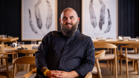 A man wearing a black shirt is sitting on a wooden chair inside a restaurant. Wooden chairs and tables are visible in the blurred background behind him. There are two large white paintings on display on the wall. The man is holding his hands in his lap. He is smiling at the camera.