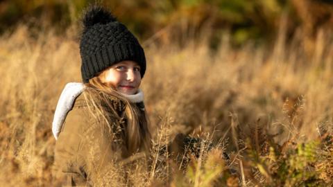 Photograph of 10-year-old Jamie, among the yellow grass. She wears a parka jacket with a fur trim, and wears a black bobble hat. She has long brown hair and blue eyes. There is an orange haze over the image. 