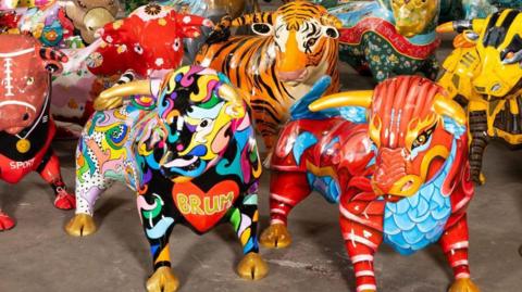 Close-up photos of a group of nearly life-size fibre-glass statues of bulls, brightly painted in various designs. The one in the foreground at the front of the picture has a red heart painted on its chest with the letters BRUM in gold inside. Another next to it is painted red and blue to look like a chinese Dragon.