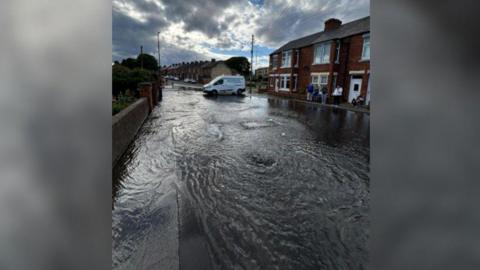 The road and the left-side pavement of Ellen Terrace covered in water, which flows down the street. A group of five people is standing outside one of the terraced houses and looking on. A white Northumbrian Water van is parked horizontally at the top of the street.
