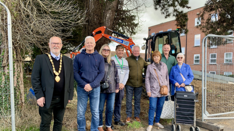 A group of council officials and residents standing at between chain link fence panels with a digger behind them