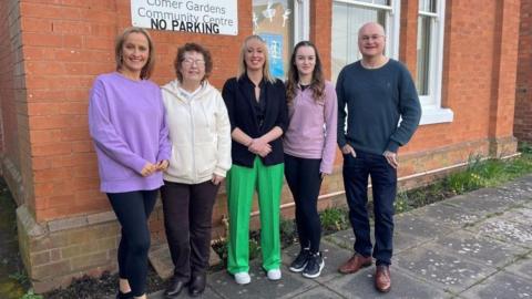 Four women and one man standing outside a red brick building with the sign "comer gardens community centre no parking" on the wall behind them.  All five smile at the camera and stand on paving slabs.