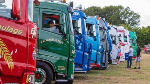 Red, Green, Blue and White HGV trucks all lined up in a row. There's several people looking at the truck parked on a green field.