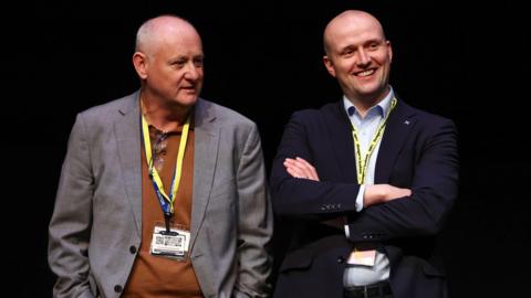 Murray Foote and Stephen Flynn - Mr Foote is wearing a grey jacket and brown shirt with a lanyard around it, while Flynn has his arms folded and is smiling