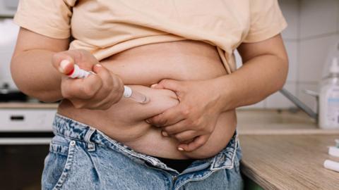 An anonymous women wearing jeans and a pale top is injecting herself in the stomach with a weight loss drug