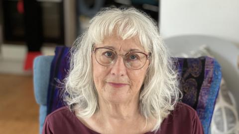 A woman wearing glasses and looking at the camera. She has blonde wavy hair with a fringe.  She is sitting down on a blue chair while wearing a burgundy full-sleeved top.