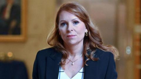 Ash Regan - a woman with long wavy red hair, wearing a dark suit and a white top -  pictured at the Scottish Parliament