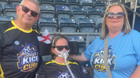 Stacy Whittall is in the middle in front of rows of blue seats at the stadium, with her father with grey and white hair on the left of the photo and her mother with brown/blonde hair on the right. They are wearing T-shirts including the words Stacy's Kick for a Cure.