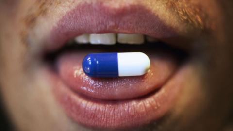 Blue and white pill sits on the tongue of a man with a moustache.