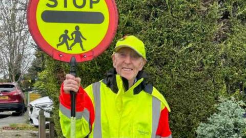 Reg dressed in high-vis uniform, coat and gate - holding a lollipop sign reading 'stop' with the highway code symbol for children/ school