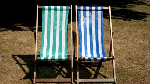 Two deckchairs placed next to eachother in a park, the one on the left is turqoise and white stripes, while the one on the right is dark blue and white stripes.