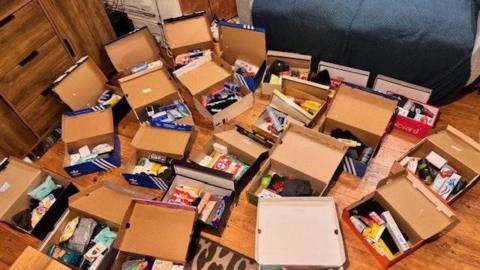 Filled shoeboxes are gathered on the floor or a room in a house. The lids are open and the contents of each box can be seen. There is a sofa and a a chest of drawers in the room, which has a wooden floor.