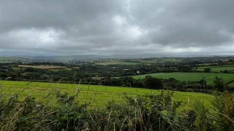 A shot of a green field ahead of a green valley full of trees and farmer's fields under a cloudy sky 