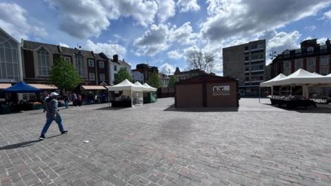A market square on a sunny day with building in the background, market stalls and a paved area.