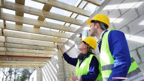 A woman wearing a hard hat has a tape measure under the beginnings of a wooden roof while a man in a hard hat stands next to her