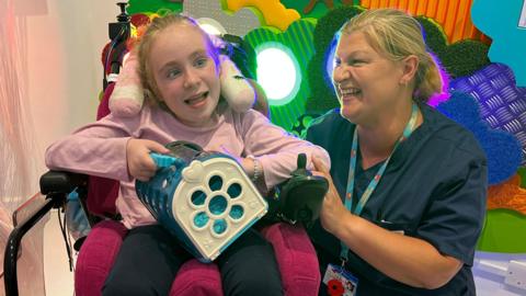 Edith Williams with Nurse Lucy Reilly in the sensory room. Edith is in a motorised wheelchair and holds a toy. Lucy is kneeling beside her, holding her arm. Both are smiling broadly and both have tied-back blonde hair.