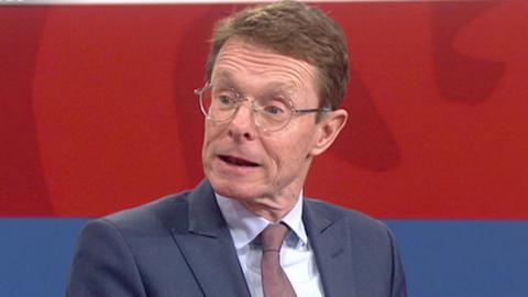 A close-up of politician Andy Street sat in a TV studio. He wears a blue suit and dark tie. His mouth is open as he is talking.