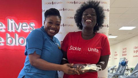 Two women at Brixton blood donation centre. One wears a blue NHS uniform and the other wears a red t-shirt that says: Sickle Cell Unite.