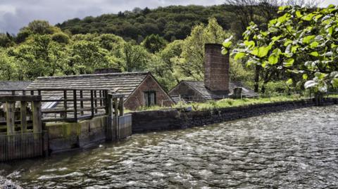 A dam with a stone wall in the foreground. Roofs of stone buildings can be seen beyond the wall.