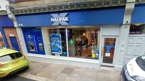 A branch of Halifax bank in Whitby. Cars are parked on the pavement outside, with people seen through the window using banking services. The bank has bold blue branding. 