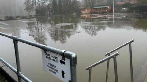 A sign which says 'club house entrance' and 'visitor changing room' in front of a completely flooded area with a brown clubhouse in the distance. The pitch is ringed by trees