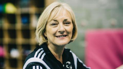 Christine is standing in a gym wearing a tracksuit. She is smiling and has bobbed blonde hair.