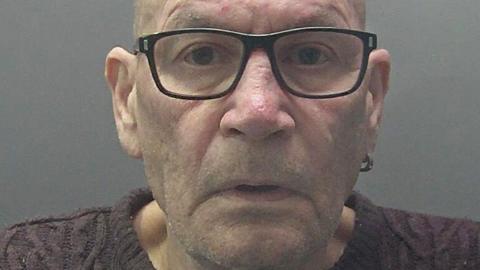 Custody photograph of David Newton, who is clean shaven and bald, looking into the camera, wearing dark glasses, a ring-shaped earring and purple jumper.