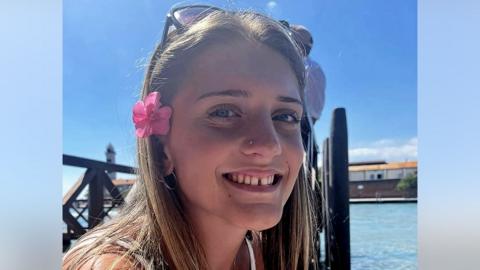 Juliette, smiling at the camera on a sunny day, is wearing her long hair down, sunglasses on her head and a pink flower tucked above her ear. She is sitting by the water