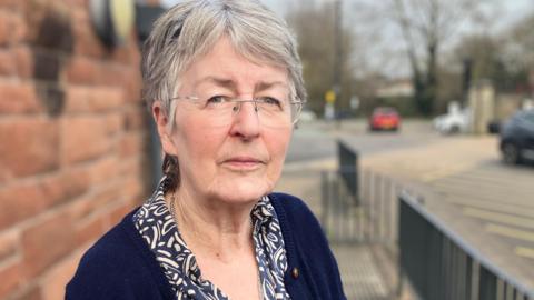 Grey-haired woman wearing rimless glasses and blue cardigan standing in between a brick wall and black railings. A car park can be seen in the background.