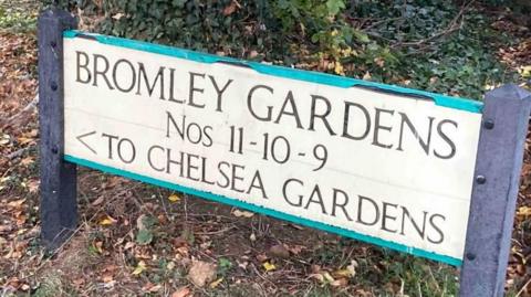 A white rectangular sign fixed to two grey posts. The words "BROMLEY GARDENS Nos 11-10-9 TO CHELSEA GARDENS" are written on the sign 