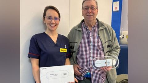 Rachel Hilmo wearing a dark blue clinical outfit holds a white cardboard box while standing with a patient, who is an older man wearing a green cardigan and a checked red and blue shirt. He is holding the wearable patch which is oval in shape and transparent on the edges and white in the centre.