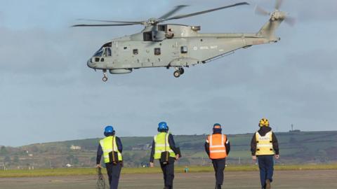 Four people are walking towards a grey Merlin helicopter which is low to the ground. They are wearing hi-viz jackets, hard hats and headphones.
