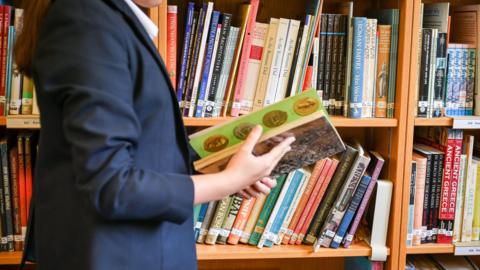 The torso of a school girl in a library, flicking through a book as she walks past a shelf full of more books.
