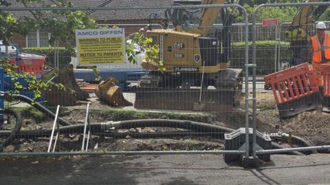 A view of construction site, behind a temporary wire fence, with black cables emerging from a hole in the road surface. A yellow digger is parked beyond the cables and a man in orange high vis uniform can be seen to the right.