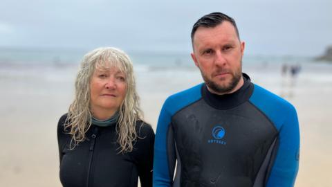 A woman and a man stand on a beach, they are wearing wetsuits. Both have wet hair and look as though they have just walked out of the sea. It is overcast and there are more people on the beach in the background