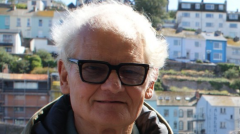 A man with grey hair and glasses stood outside in front of coloured houses. 