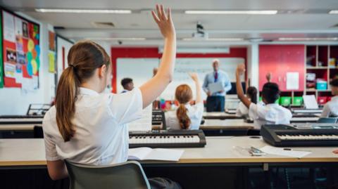 A schoolgirl wearing a white short-sleeved shirt sits at a table at the back of a classroom. She has her brown hair tied in a ponytail and there is a keyboard instrument in front of her. She has her hand in the air. In front of her are two other rows of school pupils wearing white shirts with their hands in the air. Facing them, at the front of the classroom, is a male teacher standing in front of a whiteboard.