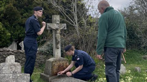 Two cadets scrub the granite memorials on a grave. Steve Davies the military grave restorer stands over them in a green fleece, giving them advice. 