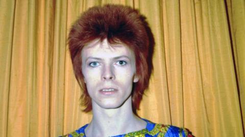 An image of David Bowie from the 1970s, he is standing against a yellow curtain and has dark red hair.