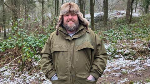 A man wearing a large green jacket and winter hat stands in a woodland, partly covered in snow.