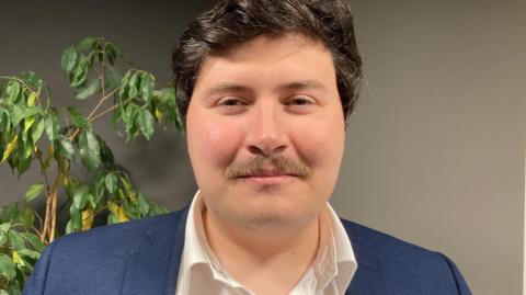 A close up of Deputy Tom Rylatt. He has short black hair, a brown moustache and is wearing a white shirt and navy jacket.