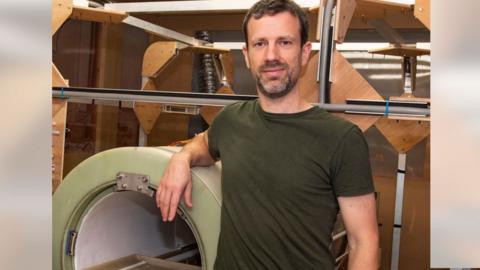 Man - Dr Lionel Broche - in green t-shirt, smiling and looking at camera, arm resting on a green scanner machine.