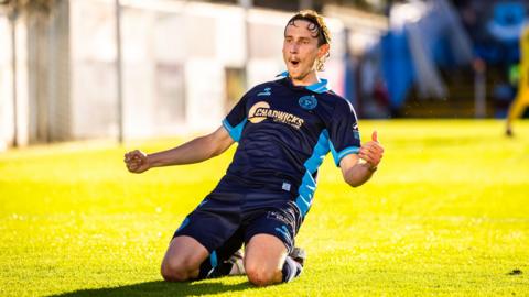 Harry Wood celebrates a goal sliding on his knees and arms outstretched, wearing Shelbourne FC dark blue kit with light blue trim