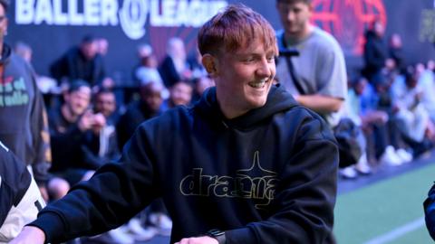 Angryginge at a Baller League event. The streamer has red hair with a long fringe and smiles while looking over his left shoulder. He wears a black hoodie and in the background, rows of people sit pitchside. 