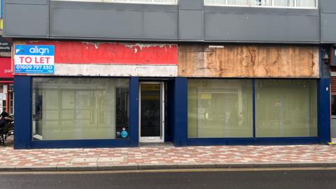 A general view of an empty commercial property in Redcar high street. The property, which gives onto a main road with double yellow lines, has a 'to let' sign above the worn-out wooden boards where shop signs would once have been. The unit, with large shop windows, is painted blue. The wooden panels are white and red on one side of the unit and rough wood on the other.