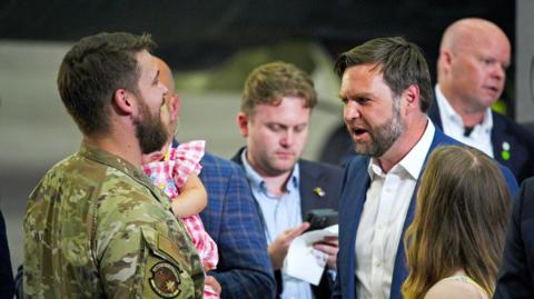 A man in military uniform is holding a young child in a pink, gingham dress in his arms. He is talking to JD Vance, who is wearing a white shirt with no tie and a blue jacket. A woman appears to be watching on, with the back of her head and her long, brown hair visible. Security personnel can be seen in the background.