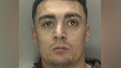 A police mugshot shows a man with short brown hair and light stubble. He is wearing a dark grey top.